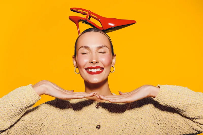 Smiling woman with closed eyes wears red high heel shoes on head and beige sweater. Bright yellow background creates cheerful mood. Concept of fashion, fun, and playful style.