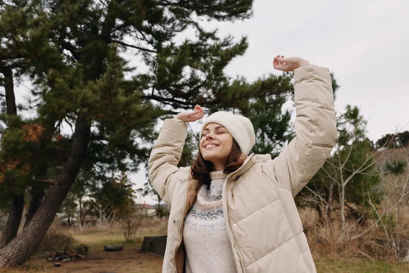 Woman outdoors in a winter jacket and beanie raises arms with a joyful smile in a park, trees and open sky in the background.