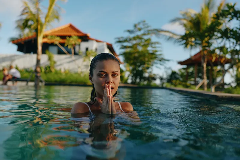Young woman practices yoga meditation in outdoor pool with tropical palm trees and villa background during sunny day, showing calm lifestyle and relaxation concept.