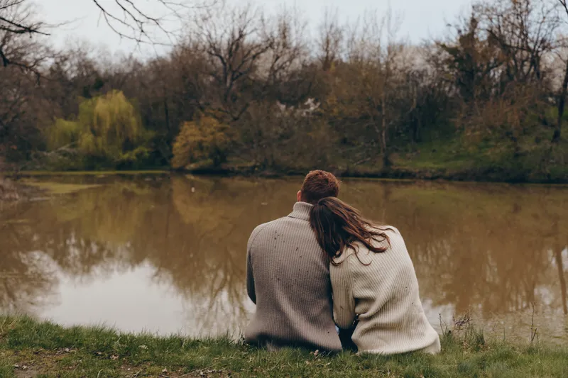 Two people sit on the grassy bank beside a calm lake, back to camera, wearing knit sweaters, sharing a quiet moment as autumn reflections shimmer on the water