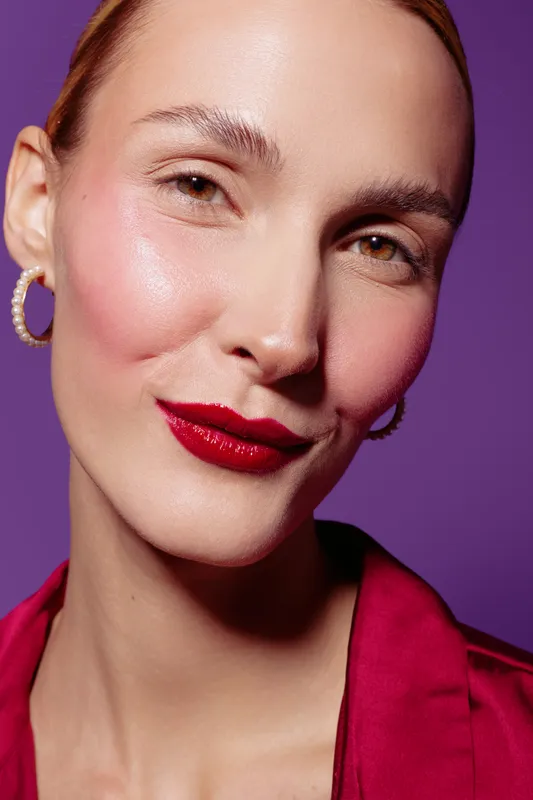 Close-up portrait of confident woman with red lipstick and pearl earrings on purple background. Concept of beauty, fashion, makeup, and elegance with soft studio lighting.