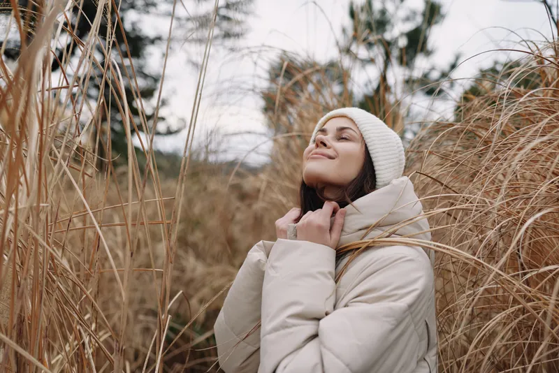 A woman outdoors in a winter coat and scarf, wearing a beanie, enjoying nature and a peaceful pose among tall grasses, capturing a quiet winter mood outdoors
