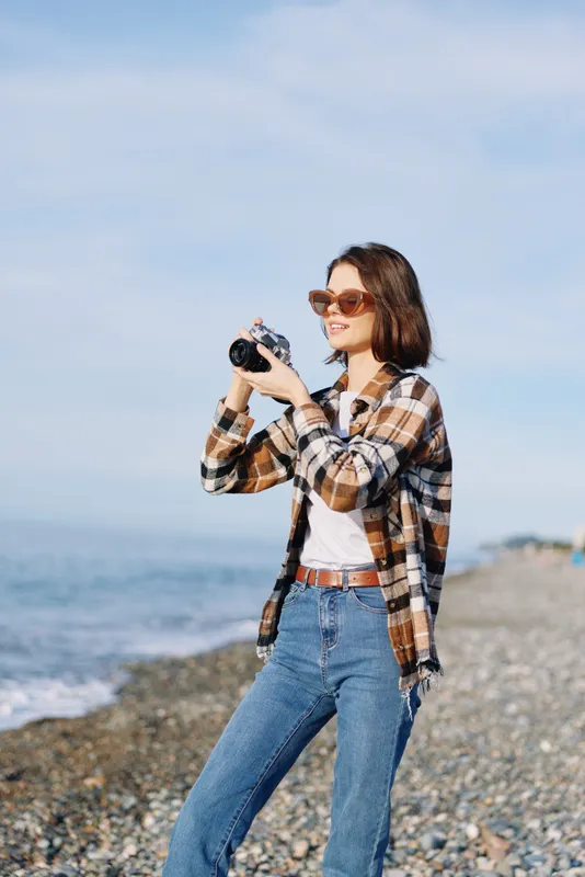Photographer woman on a windy beach holds a camera, wearing sunglasses and a plaid shirt, jeans and a belt, capturing seaside scenery and candid moments along the coastline.