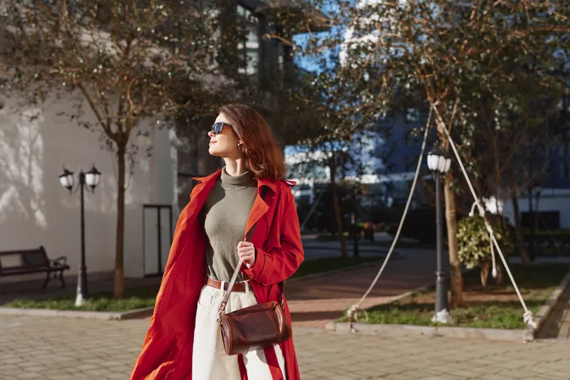 A confident woman in a red coat and sunglasses walks outdoors on a sunny day, carrying a brown handbag. Autumn trees and urban street lamps are visible in the background.