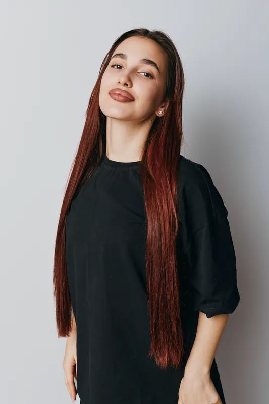 Woman with long straight hair wearing black dress standing against plain background looking confident and relaxed with natural makeup and earrings in studio portrait.