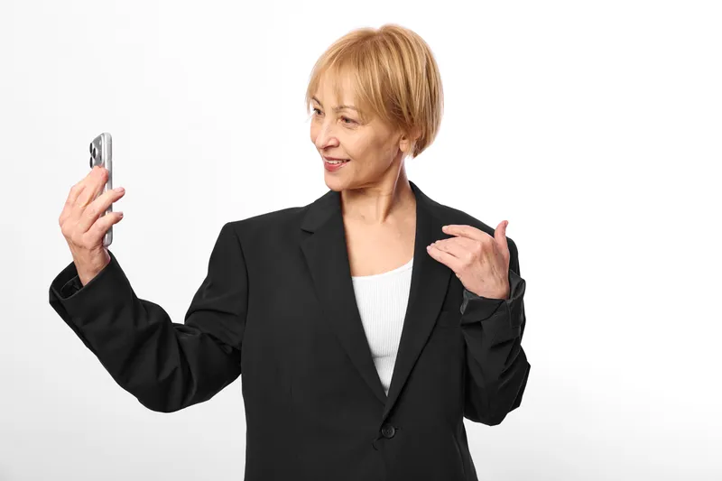 Confident business woman in black blazer gestures while holding a smartphone, using technology for communication on a clean white background, portrait concept.