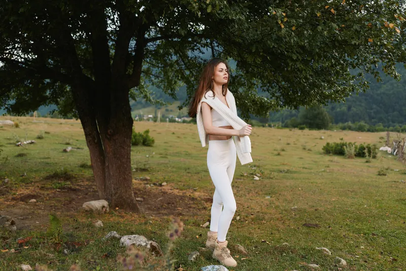 Young woman in white outfit standing near a tree in a green meadow. Outdoor lifestyle scene with nature and calm mood during daytime.