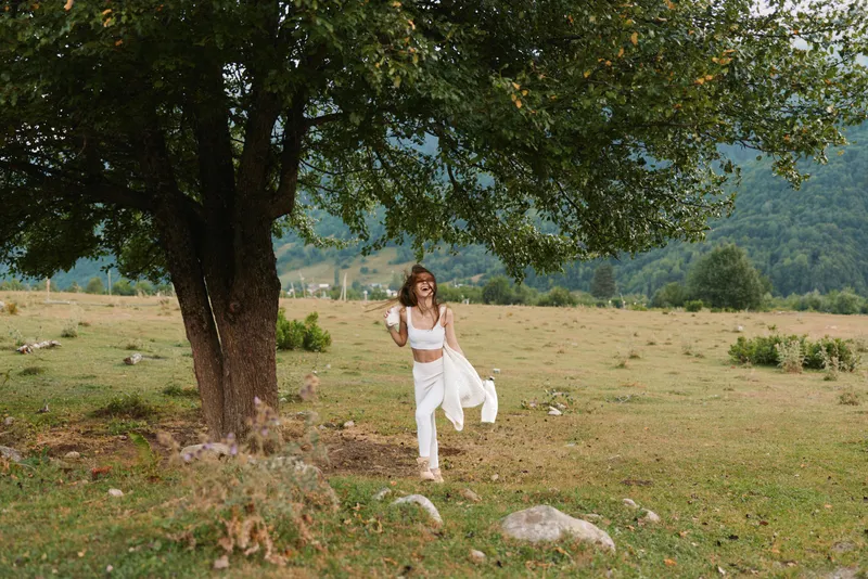 Woman in a white flowing dress walks through a grassy field under a large tree, enjoying a sunny day, conveying carefree outdoor lifestyle, natural beauty and graceful movement and freedom