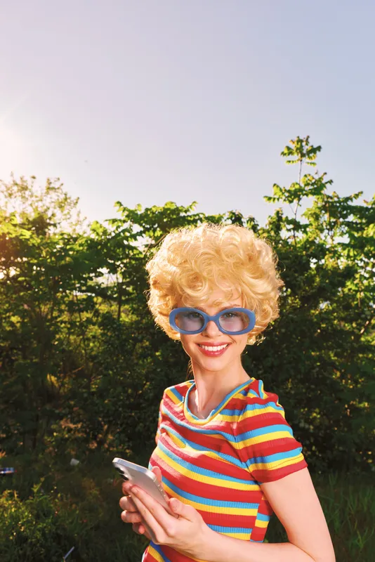 Retro blonde woman with a curly wig wears a rainbow striped shirt and blue sunglasses outdoors, smiling at the camera under bright daylight in a sunny garden setting with green trees behind.