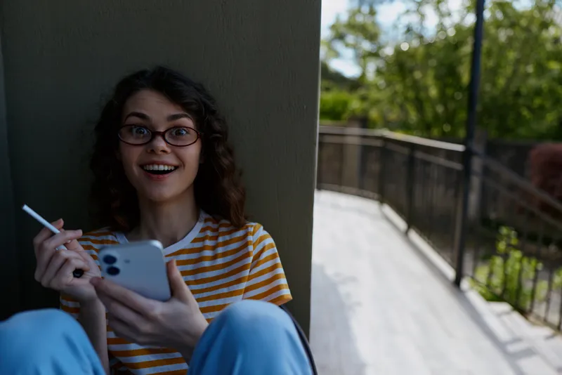 A cheerful young woman with glasses sits on a balcony enjoying outdoor light, holding a smartphone in one hand and a cigarette in the other. Casual lifestyle portrait, relaxed youth moment.