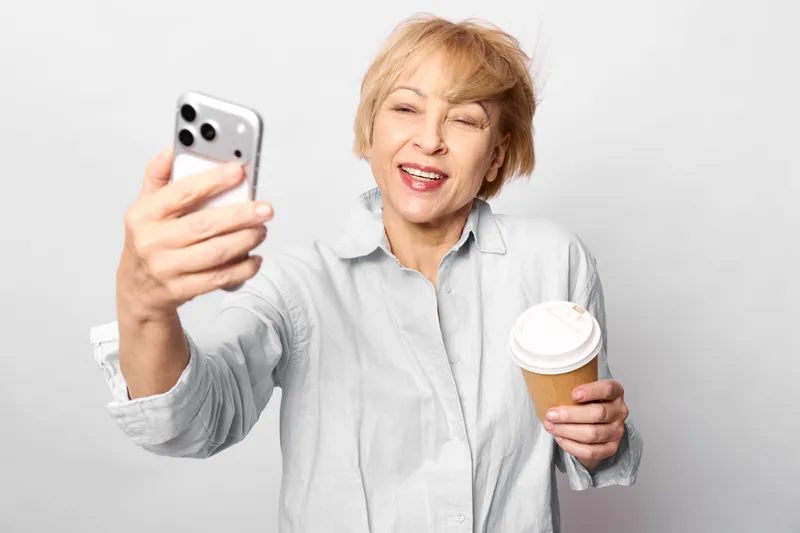 Elderly woman portrait, holding smartphone for selfie, coffee cup in hand, smiling with closed eyes, casual indoors, bright white background