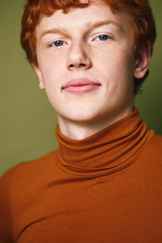 Portrait of young man with red hair and fair skin wearing brown turtleneck, looking confidently at camera on green background with natural lighting in studio.