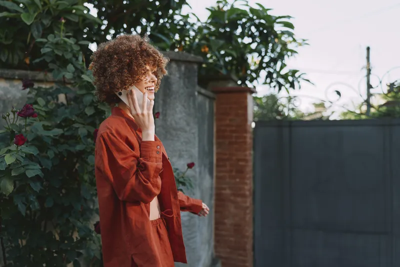 A stylish young woman with curly hair in an orange coat walks along a sunlit alley by a garden wall, talking on her phone and smiling warmly at a casual moment