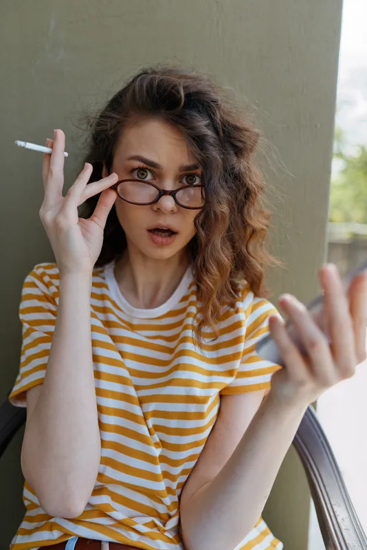 Young woman with curly hair wearing a striped yellow shirt and glasses, indoors, surprised expression, cigarette in hand, casual lifestyle moment at home, relaxed pose and natural light contribute to