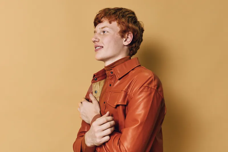 Redhead young man wearing orange leather jacket posing on beige background with confident expression, casual style and side profile portrait in studio light.