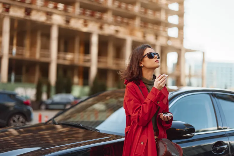 A stylish woman in a red coat stands beside a sleek black car on an urban street, adjusting her sunglasses as she gazes into the distance.