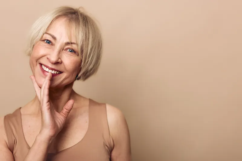 Smiling mature woman with short blonde hair in beige top touches her face gently, posing against plain neutral background. Concept for beauty, confidence, and natural aging.