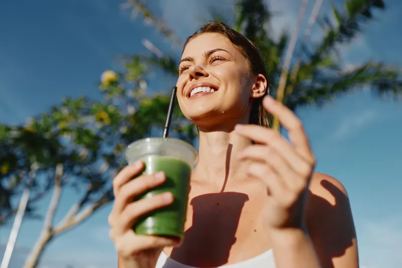 Smiling woman in a sunny outdoor setting holds a green smoothie cup with a straw, enjoying a refreshing healthy moment. Bright sky and tropical trees convey wellness, vitality, and self care.