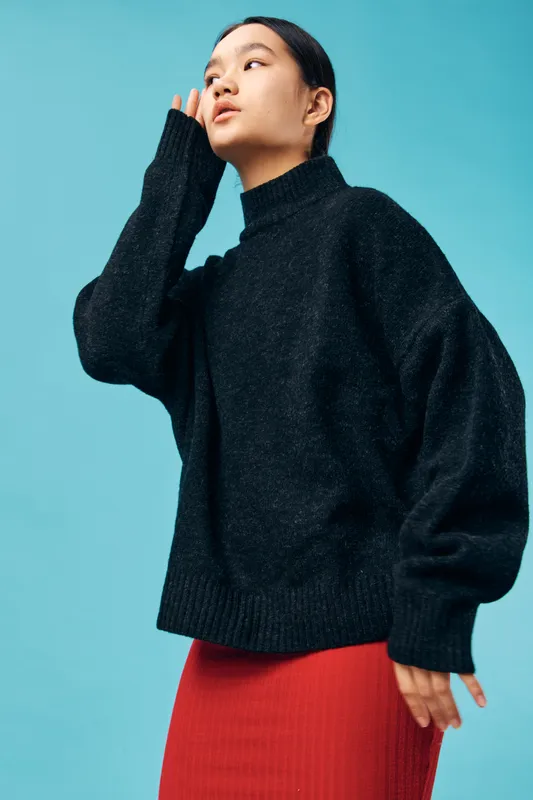 Young woman with black sweater and red skirt posing against blue background with hand near face in stylish casual clothing for fashion and portrait photography with confident expression.