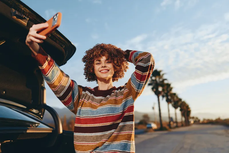 Woman lifestyle smartphone car road outdoors smiling curly striped travel, young female in striped sweater holds smartphone near open car trunk, sunny street bokeh, casual vacation mood
