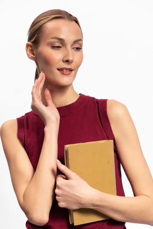 Young woman in sleeveless red top holds a closed book, looking aside with a slight smile. Studio portrait on white background. Concept of reading, education, and thoughtful mood.