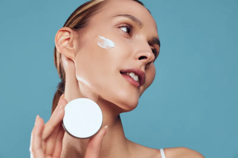 Young woman with clear skin applies face cream on her cheek while holding a white cosmetic jar against a blue background, showing skincare routine and fresh natural look.