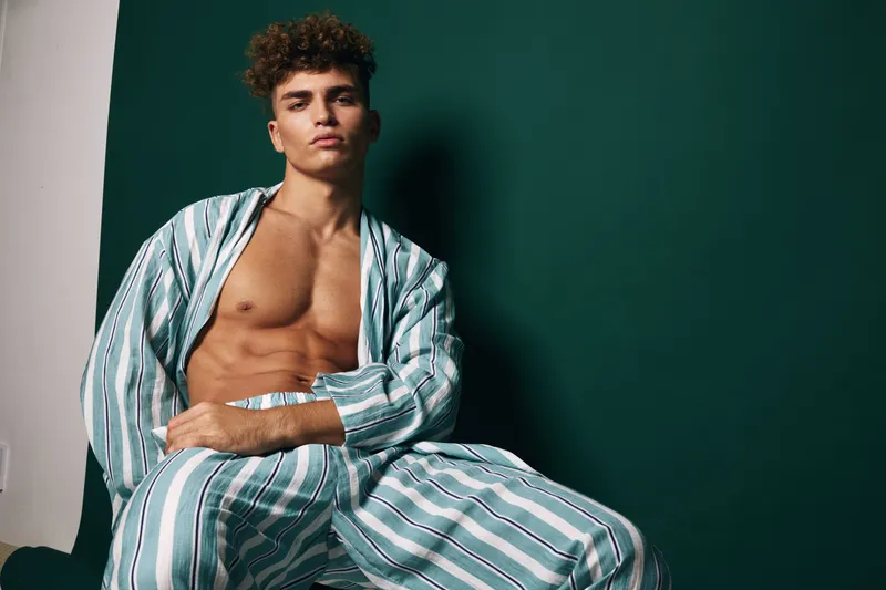 Young man with curly hair wearing striped pajamas sitting against dark green wall in relaxed pose. Casual fashion, male model, indoor portrait with confident expression and soft lighting.