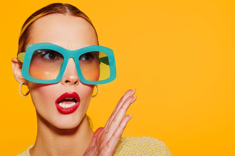 Stylish woman with large blue sunglasses and red lipstick poses with surprised expression on bright yellow background. Concept of fashion, beauty, and summer style.