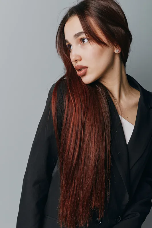 Young woman with long straight brown hair wearing black blazer and white top posing looking sideways on gray background. Fashion model with natural makeup and earrings in studio portrait.