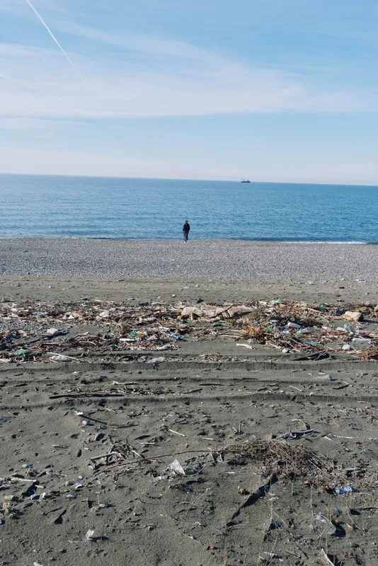 Beach scene with scattered litter along the shoreline, a lone figure gazing at the calm sea under a blue sky, highlighting waste and environmental concerns.