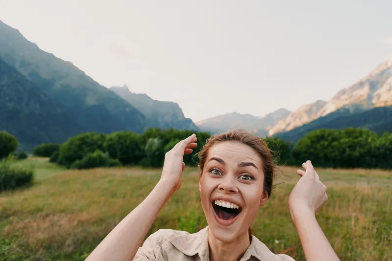 Happy woman outdoors in nature, smiling widely with raised hands, mountain landscape background, casual clothing, bright daylight, joyful expression, fresh air