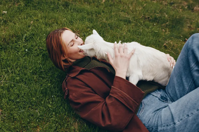 A woman in a brown coat lies on grass, sharing a gentle kiss with a white goat kid, capturing an outdoor lifestyle moment of animal affection and connection in bright daylight