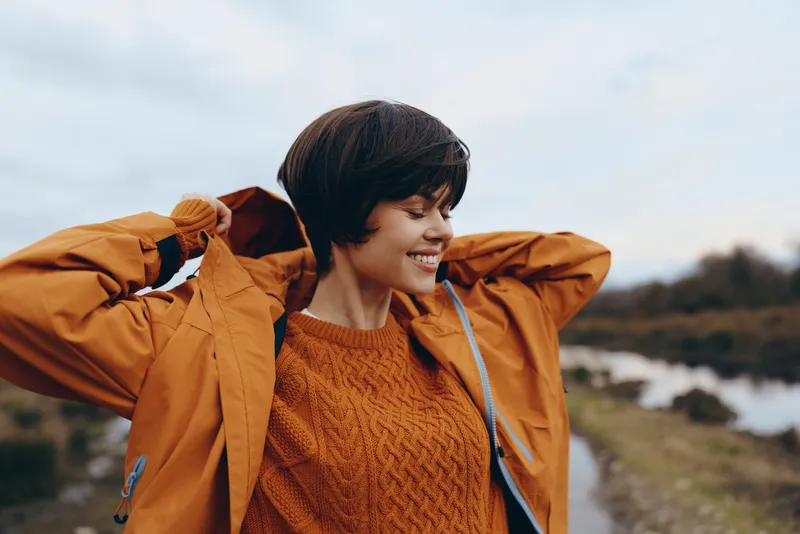 Woman outdoors lifestyle, smiling traveler in orange sweater and jacket, holding backpack straps, gentle breeze, walking countryside path, near river, soft daylight, autumn mood