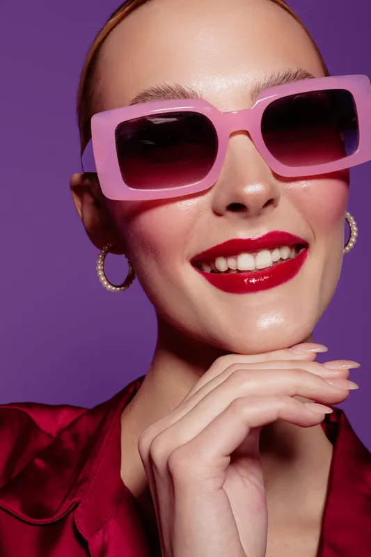 Smiling woman with pink sunglasses and red lipstick poses against purple background. Close-up portrait with hoop earrings and shiny skin. Concept of fashion, beauty, and style.