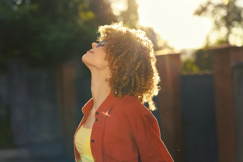 A woman in casual outfit stands outdoors in bright sunlight, head tilted back, a relaxed hopeful moment captured in daytime light for a lifestyle portrait