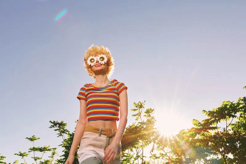 Woman lifestyle portrait outdoors in a striped crop top and light shorts, retro sunglasses with flower frames, sunny day vibe, casual playful mood, vibrant colors in natural light.