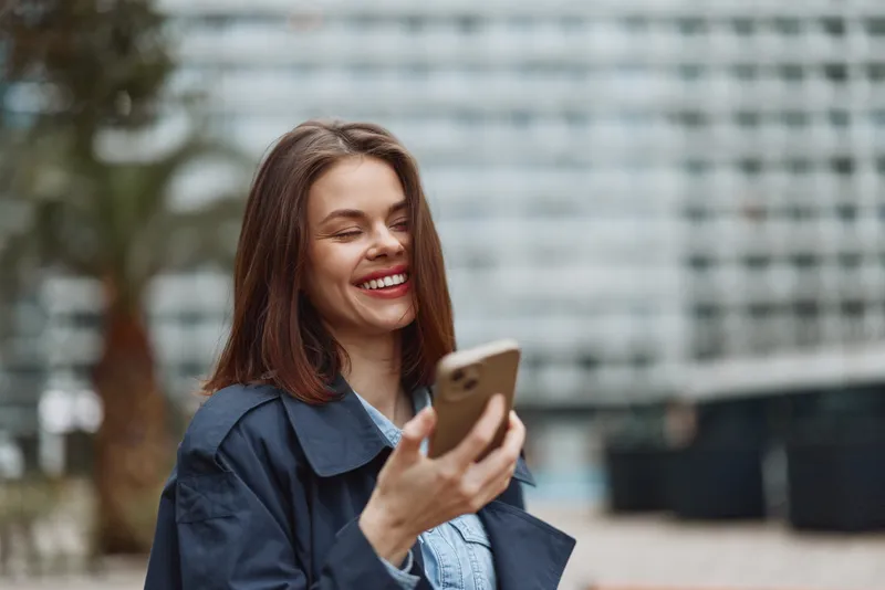 A smiling young woman in a dark coat holds a smartphone outdoors, looking away from the camera. The blurred urban background suggests a casual and happy moment.