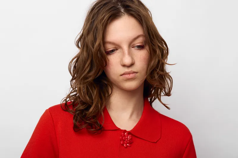 Sad young woman with curly brown hair wearing red sweater looks down with thoughtful expression on white background. Concept of loneliness, contemplation, and emotional struggle.