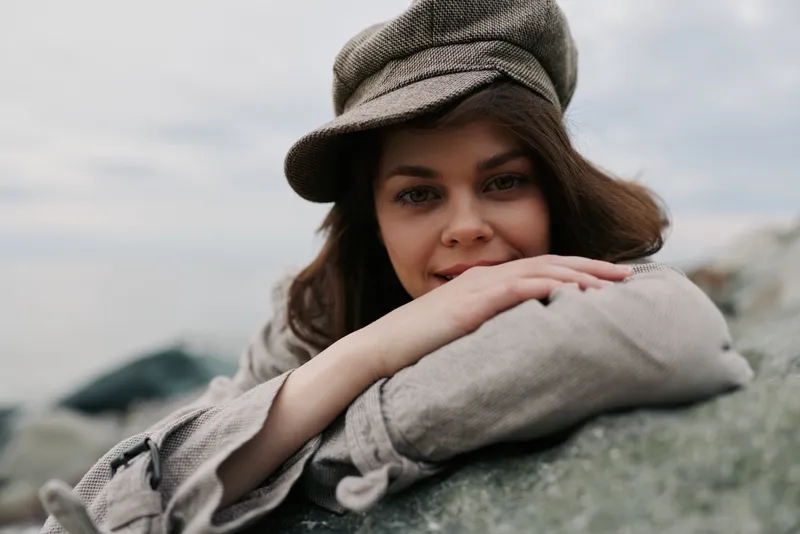 Young woman in a stylish hat lies resting on a rock outdoors, smiling gently with soft natural light. Concept for relaxation, nature, and casual fashion portrait.