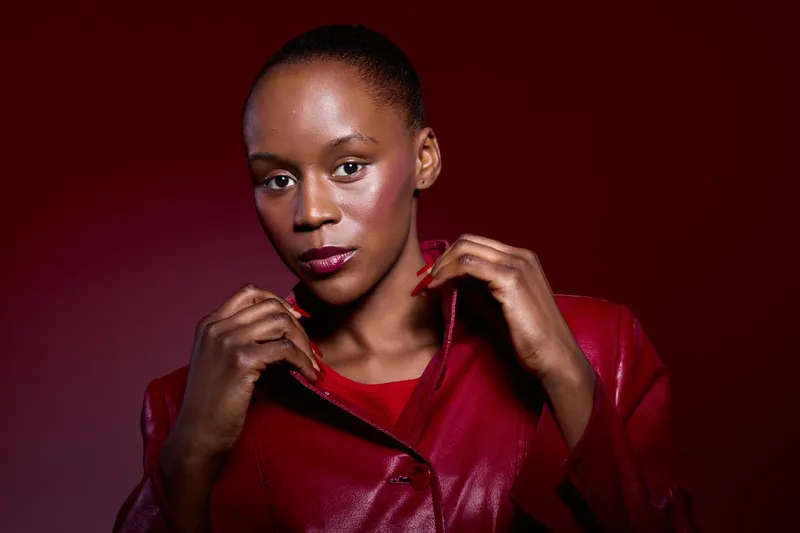 Confident African woman with dark skin wearing a red leather jacket poses against a dark red background. Concept of fashion, beauty, and empowerment in studio lighting.