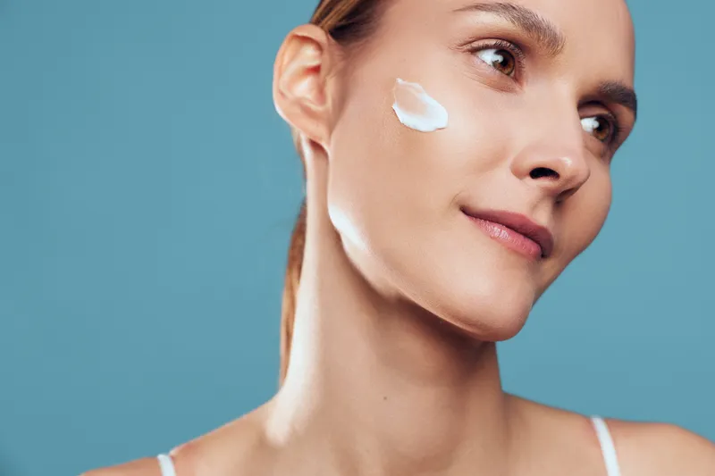 Young woman applies facial cream on her cheek during a skincare routine, isolated on a blue background with calm mood and a gentle diffuse lighting enhances the clean beauty shot.