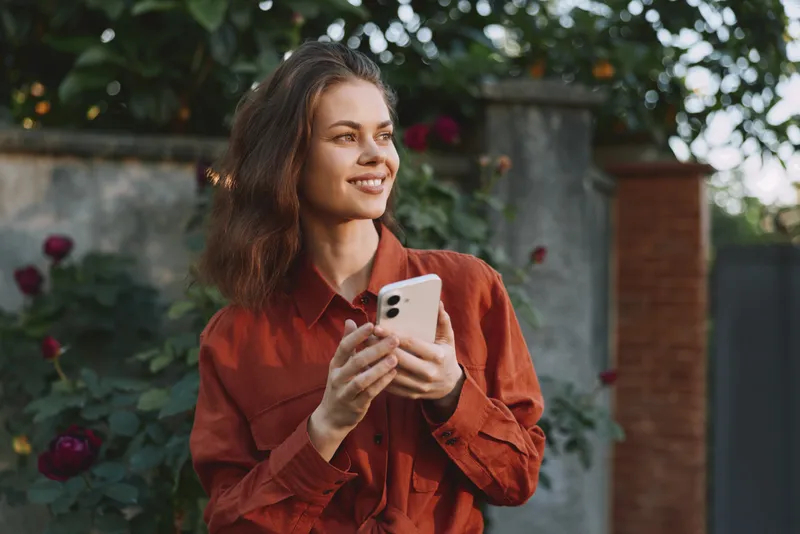 Woman lifestyle outdoor scene, smiling, holding a phone, wearing a rust shirt, standing in a garden with flowers, conveying casual fashion and natural confidence in daylight