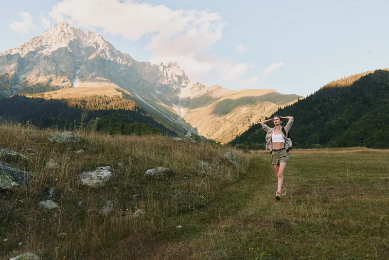 A woman hiker stands in a sunlit meadow with mountains behind, backpack on shoulders. A lifestyle moment of outdoor exploration and nature.
