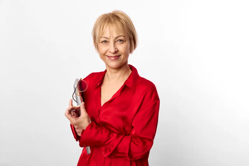 Senior woman in red robe holding glasses, smiling in studio portrait on clean white background, warm caring expression, soft light, minimal backdrop, relaxed friendly mood