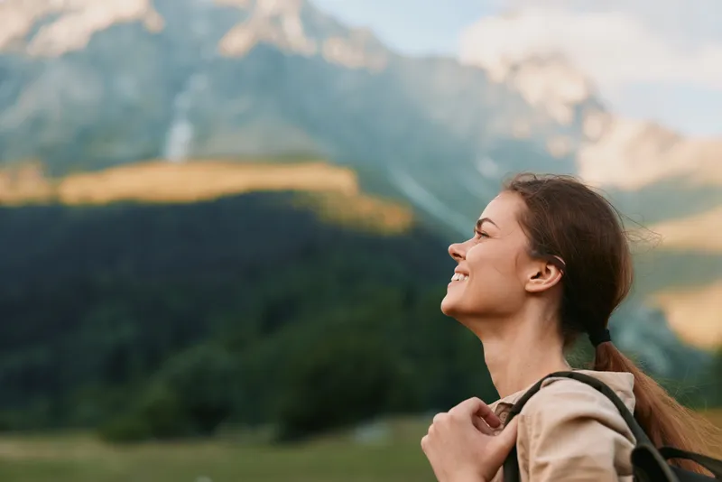 Young woman with long brown hair smiles happily while hiking in the mountains during a sunny day, enjoying nature and carrying a backpack on her shoulders.