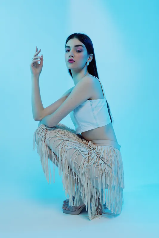 Young woman beauty fashion model poses in fringed pants and white top against blue background. Studio shot emphasizing style, elegance, and modern trends in clothing and makeup.