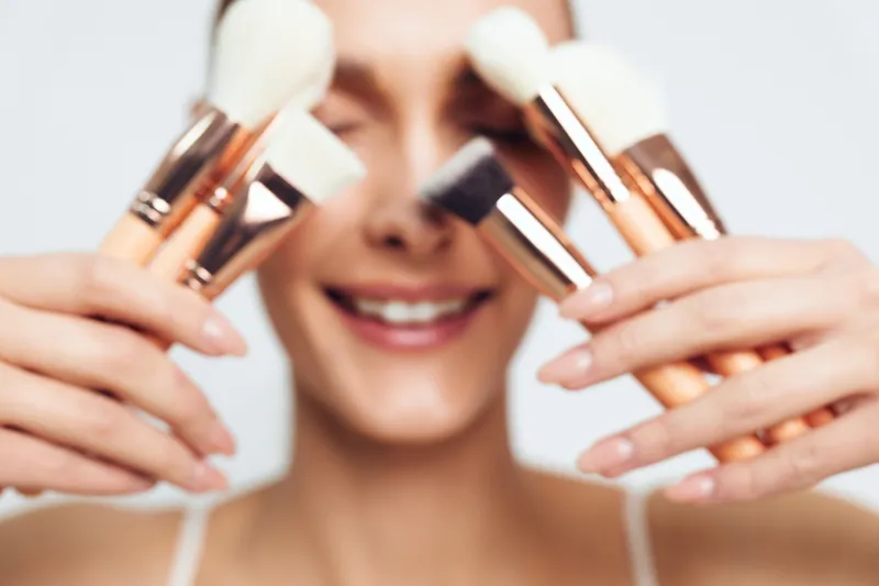 Cheerful woman holding makeup brushes, closeup portrait, smiling with soft light on skin, beauty tools ready, subtle makeup tones, natural glow and relaxed vibe, indoor studio