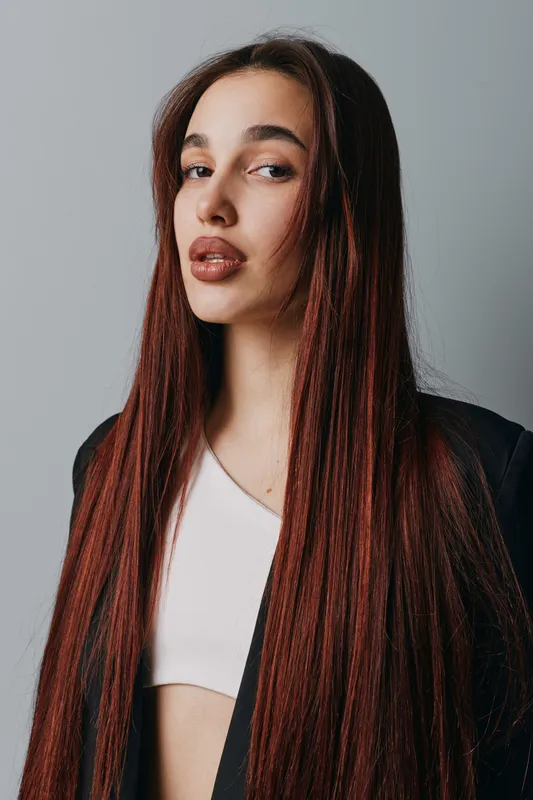 Woman with long straight hair and natural makeup posing in white top and black jacket against gray background. Portrait of confident young female with serious expression and stylish look.