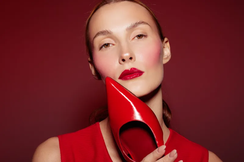 Close-up of a woman with red lipstick holding a shiny red high heel shoe near her face. Red background and red clothing create a bold fashion and beauty concept with elegant style.