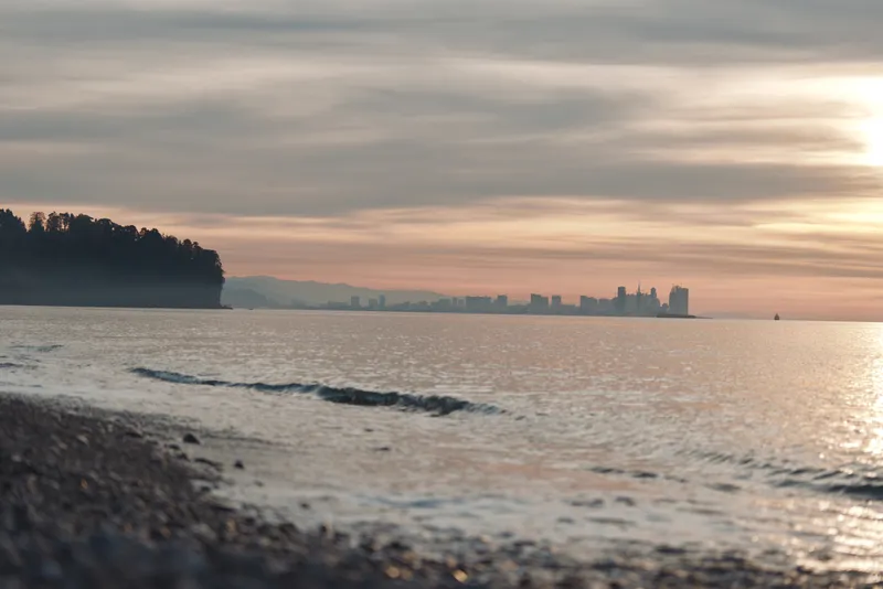 Beach and calm shore at sunset over the water with a distant city skyline, gentle waves, warm golden light and a serene horizon creating a tranquil coastal landscape.
